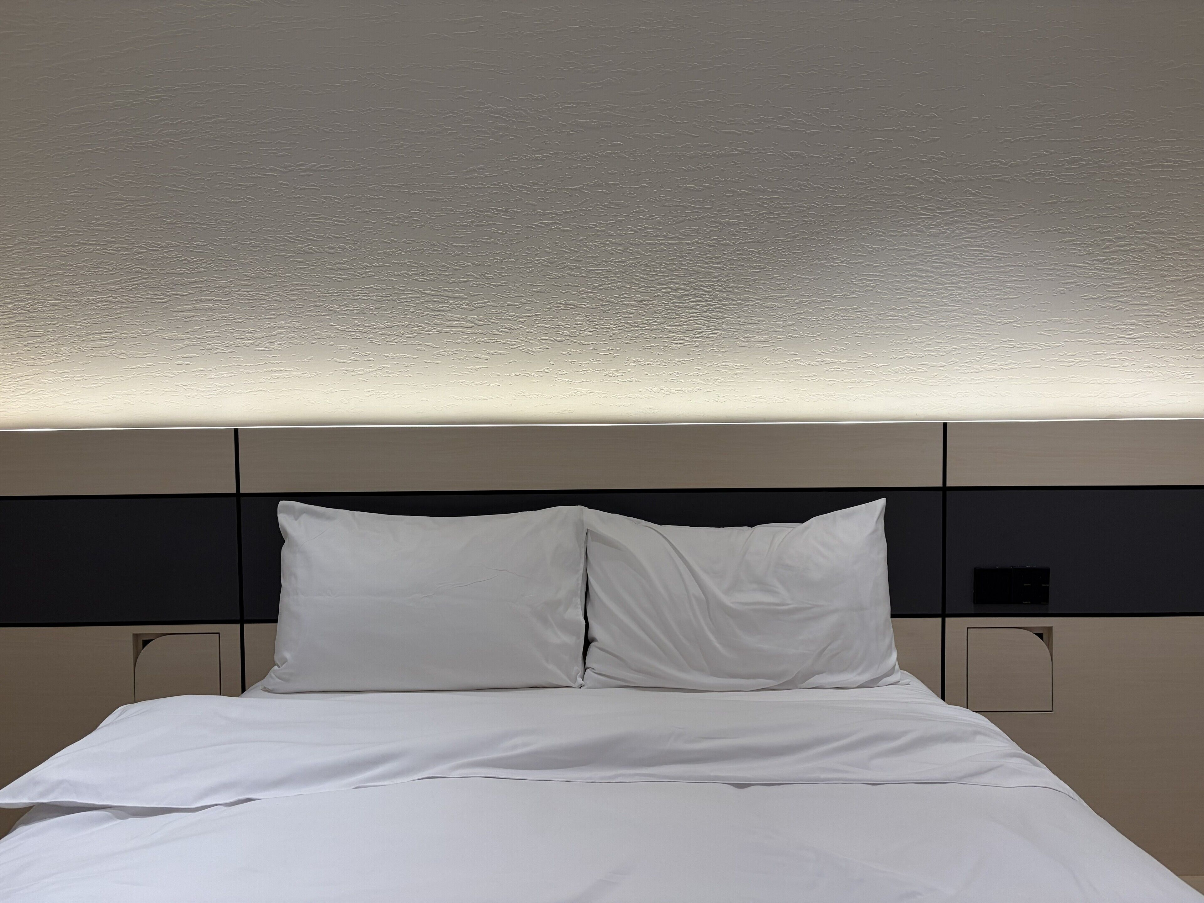 A hotel room with a bed, white bedding, and a headboard with storage compartments.
