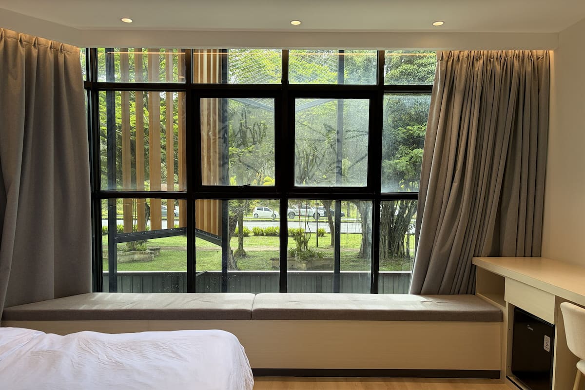 A bedroom with a large window, a bed, a desk, and a bench.