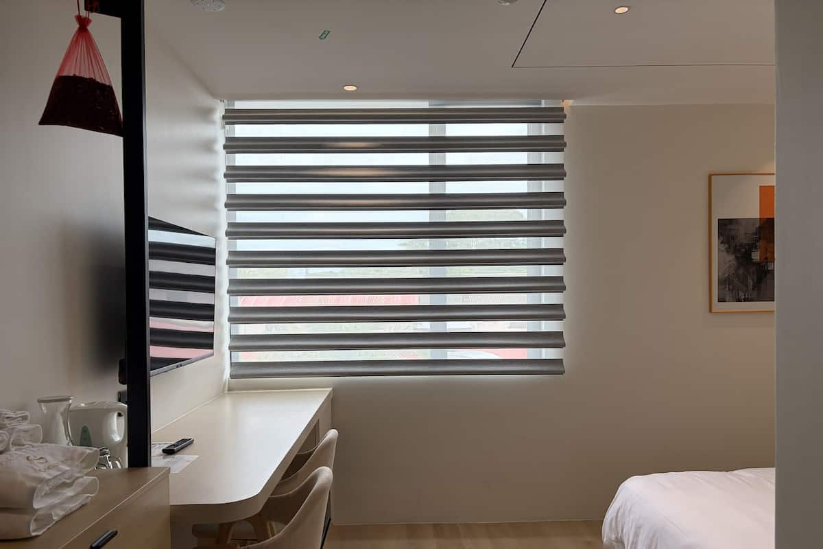 A modern hotel room with a large window, a bed, a desk with chairs, and a small table with a hairdryer and towels.