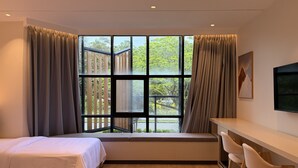 A modern hotel room with a large window, a bed, a desk with chairs, and a mounted TV.