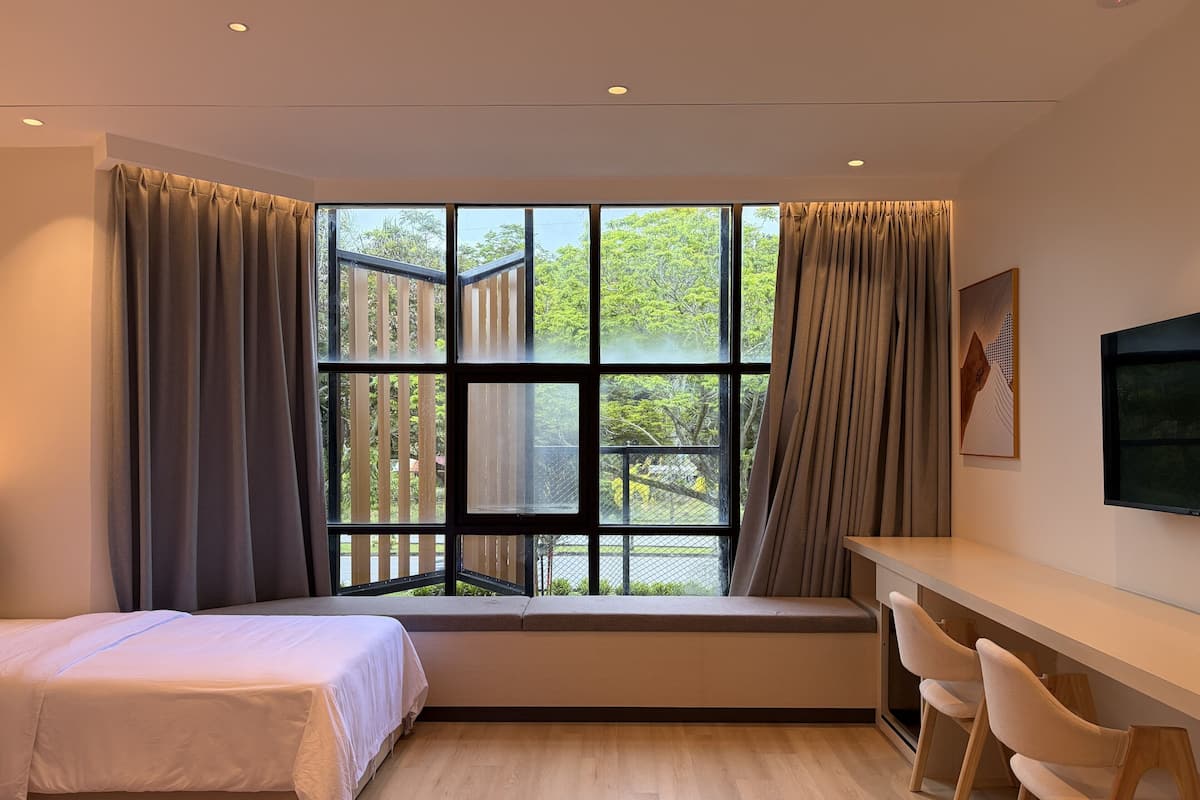 A modern hotel room with a large window, a bed, a desk with chairs, and a mounted TV.