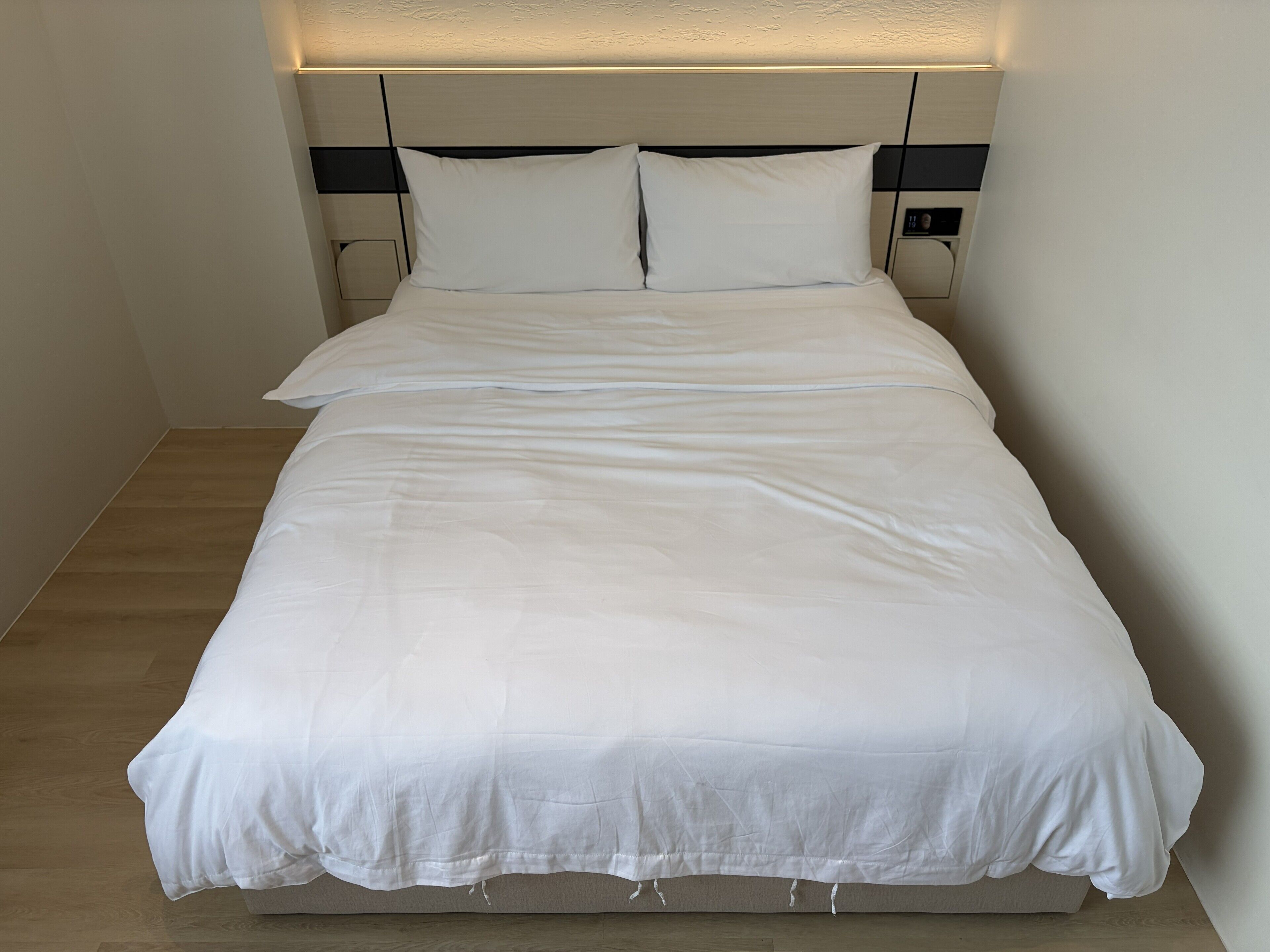 Een net opgemaakt bed met witte lakens, een hoofdbord met lichtschakelaar en een houten vloer.