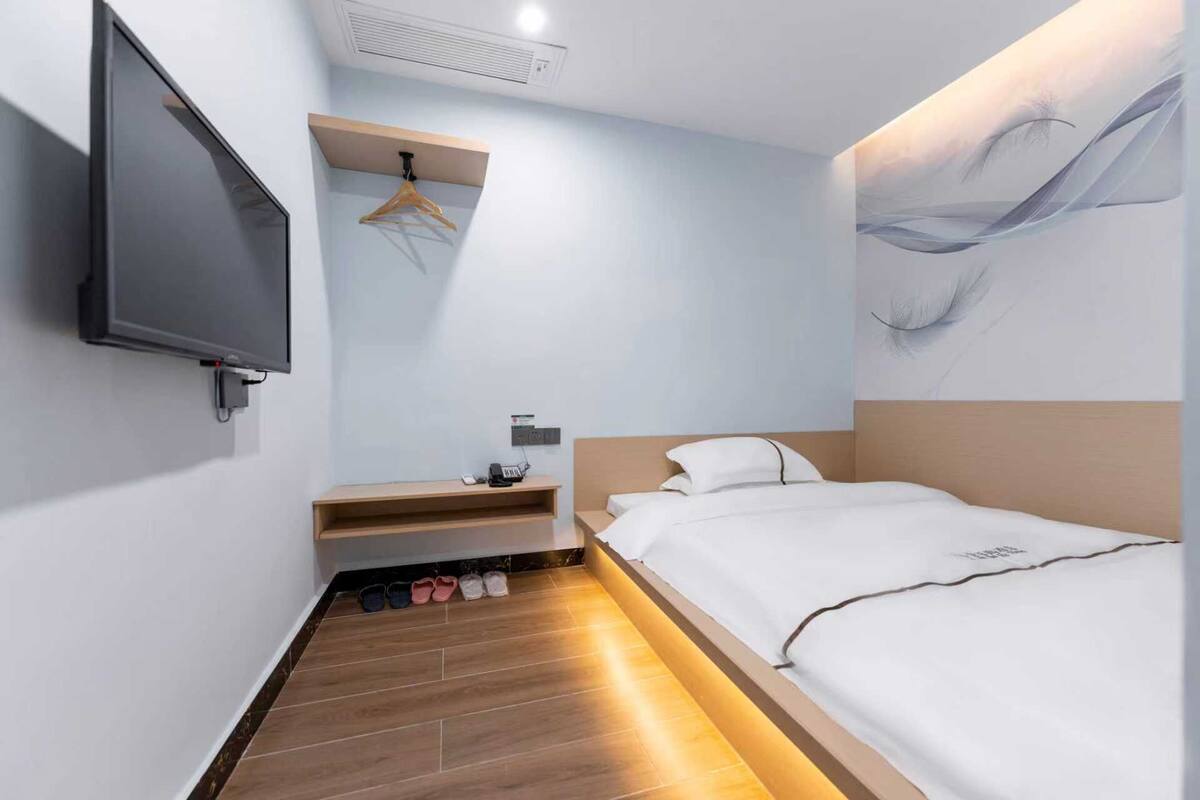 Ein modernes Hotelzimmer mit einem Bett, einem an der Wand befestigten Fernseher, einem Schuhregal und einem kleinen Ablagebrett mit Telefon und Ladegerät.