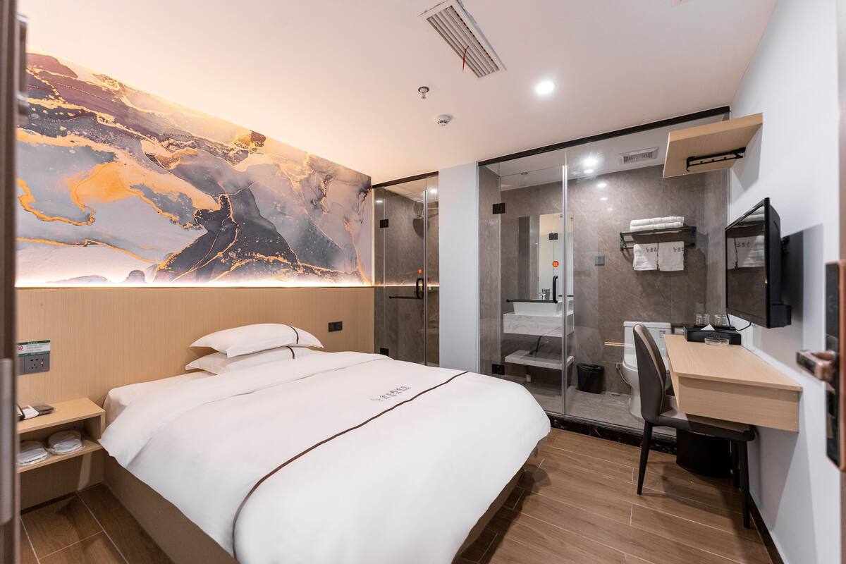 Ein Hotelzimmer mit Bett, Schreibtisch, Duschbereich und einem großen, abstrakten Gemälde an der Wand.