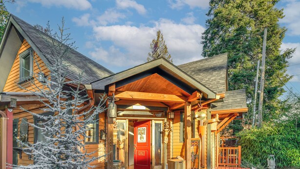 Une cabane en bois avec une porte rouge, un porche abrité et un arbre recouvert de neige devant.