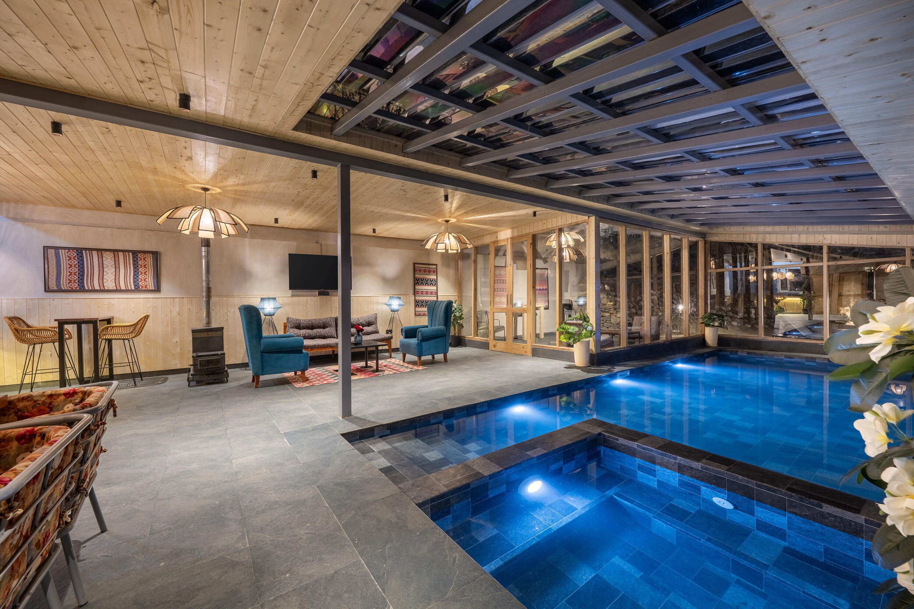 Un área de piscina interior moderna con techo de madera, paredes de vidrio y una zona de estar con sillas azules.