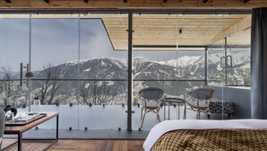 A modern room with a large glass window, a bed, a table with a chair, and a view of snow-capped mountains. - Echor Himalayan Sky Cabins Sethan Manali (Manali)