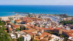 Paisagem costeira com edifícios históricos, um porto de iates e um cais que se estende até o mar.