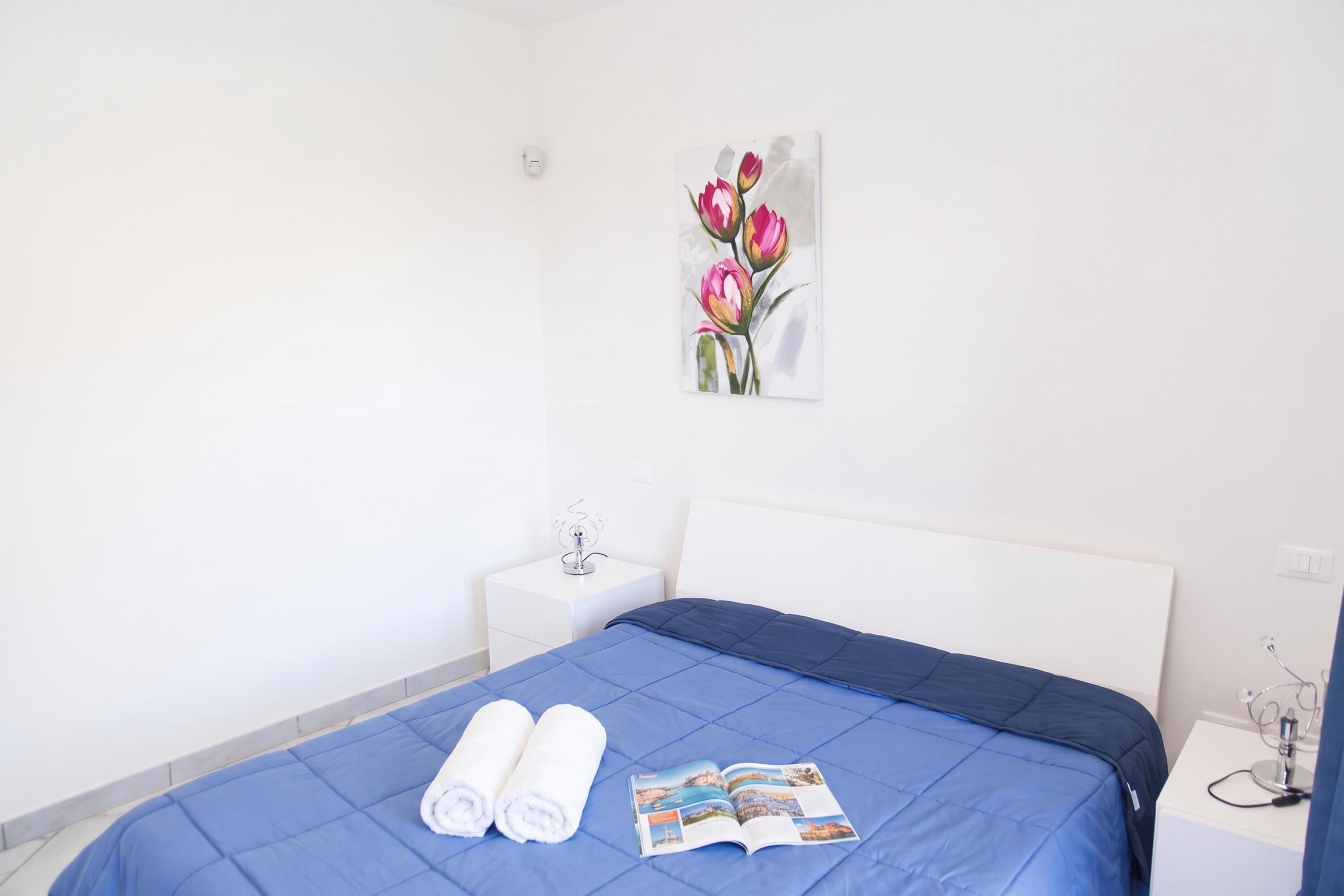 Una habitación de hotel con una cama, una mesita de noche, un interruptor de luz en la pared y un cuadro de flores.