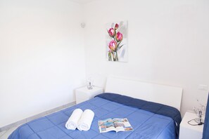 A hotel room with a bed, a nightstand, a wall-mounted light switch, and a painting of flowers. - Apartments in Villa Fortunata (Terracina)