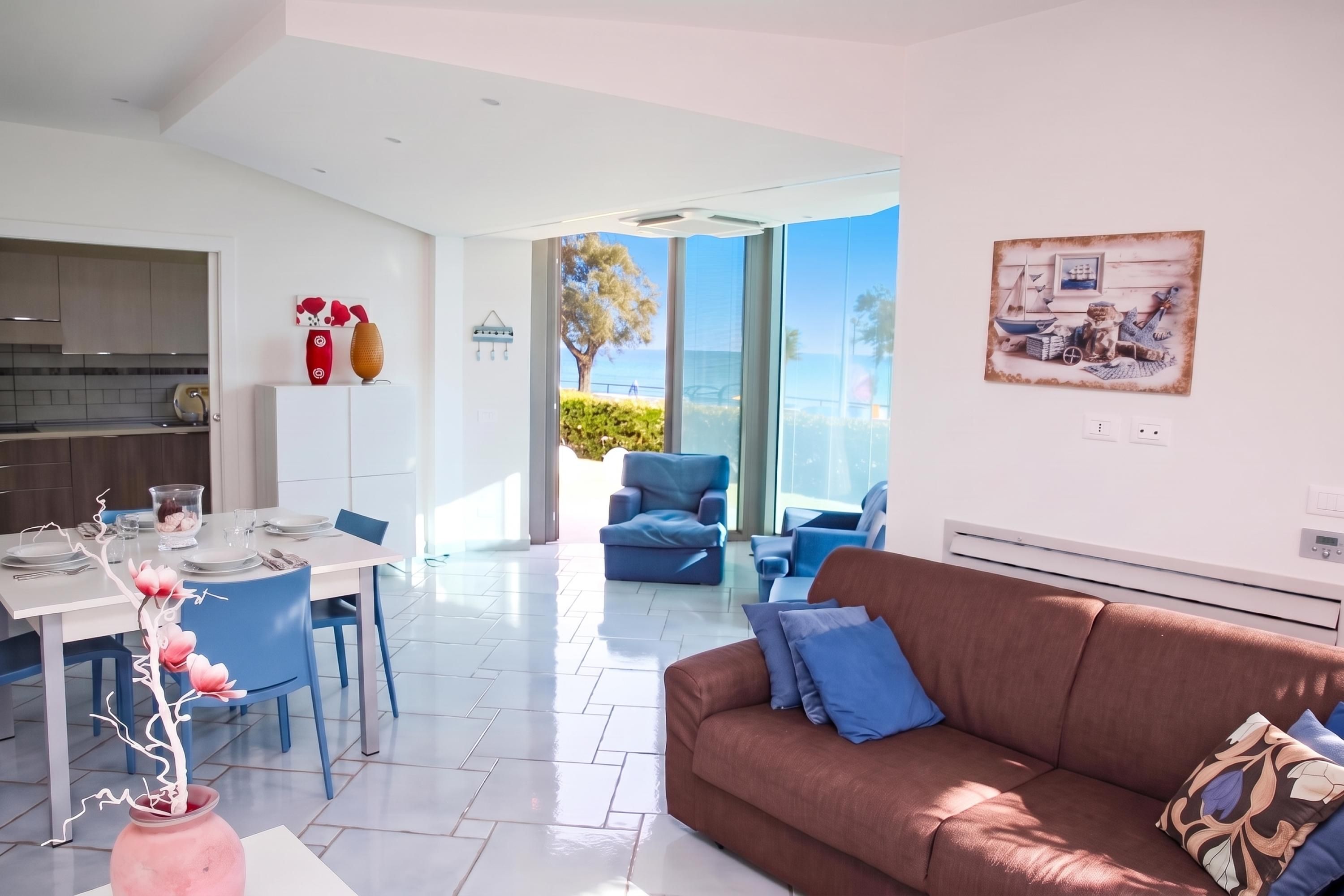 Una sala de estar moderna con un sofá marrón, una zona de comedor con una mesa blanca y una cocina con armarios de madera.