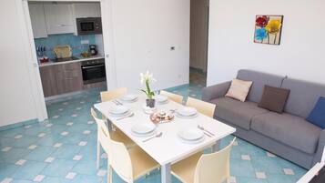 Uma sala de jantar com mesa posta para quatro pessoas, um sofá e cozinha ao fundo.