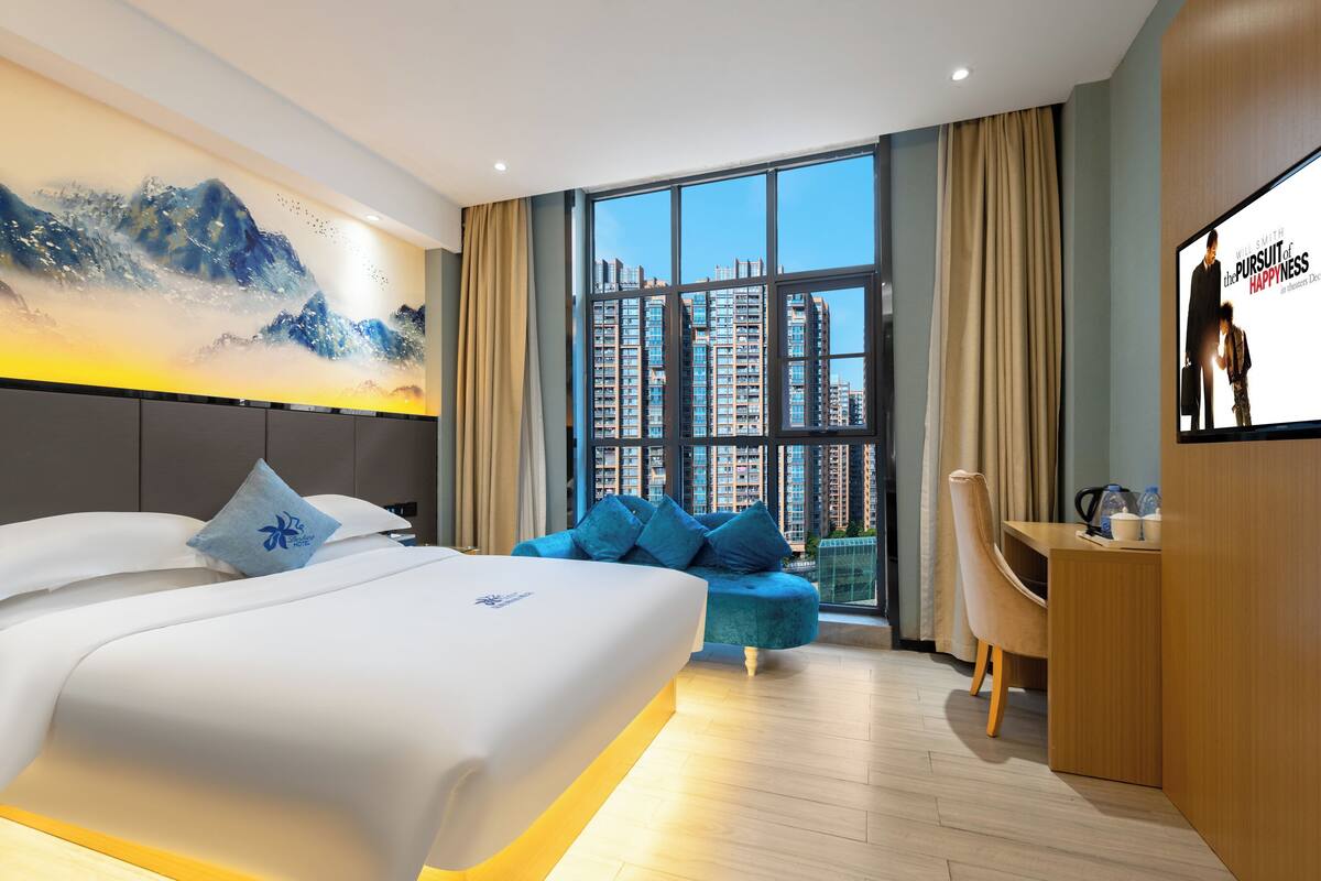 Ein modernes Hotelzimmer mit einem großen Bett, einem Schreibtisch, einer Sitzecke und einem großen Fenster mit Blick auf die Stadt.