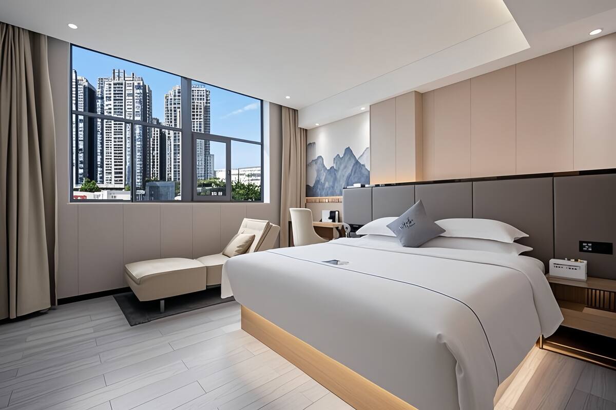Ein modernes Hotelzimmer mit einem großen Bett, einem Schreibtisch, einem Stuhl und Blick auf hohe Gebäude.