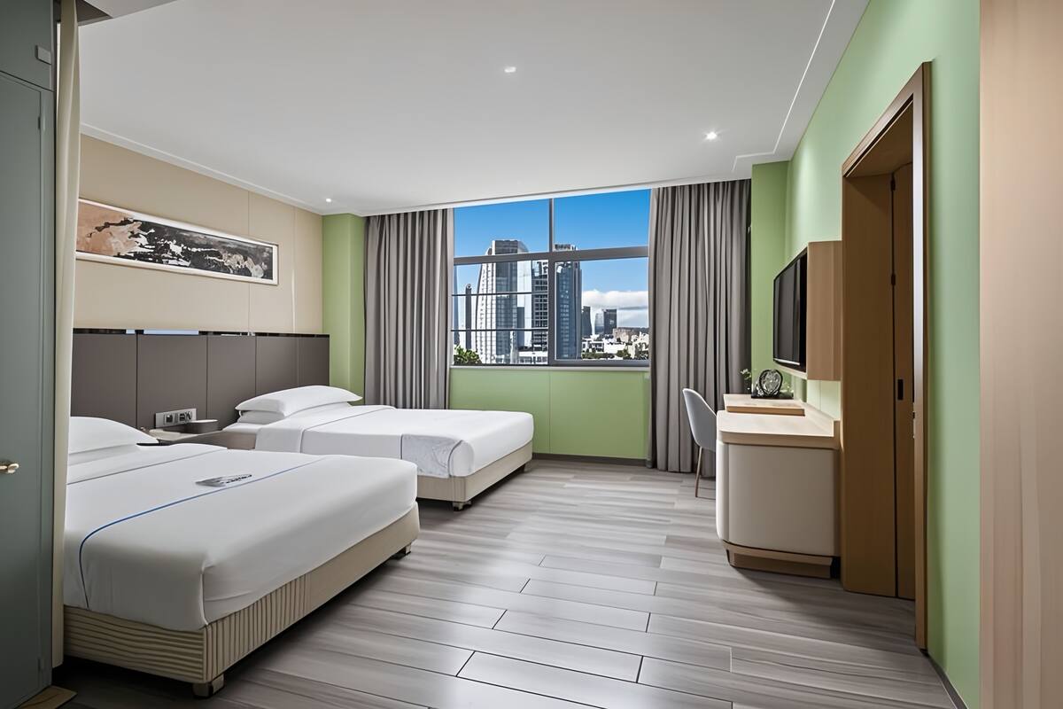 Ein Hotelzimmer mit zwei Betten, einem Schreibtisch, einem Sessel, einem Fernseher und einem großen Fenster mit Blick auf die Stadt.