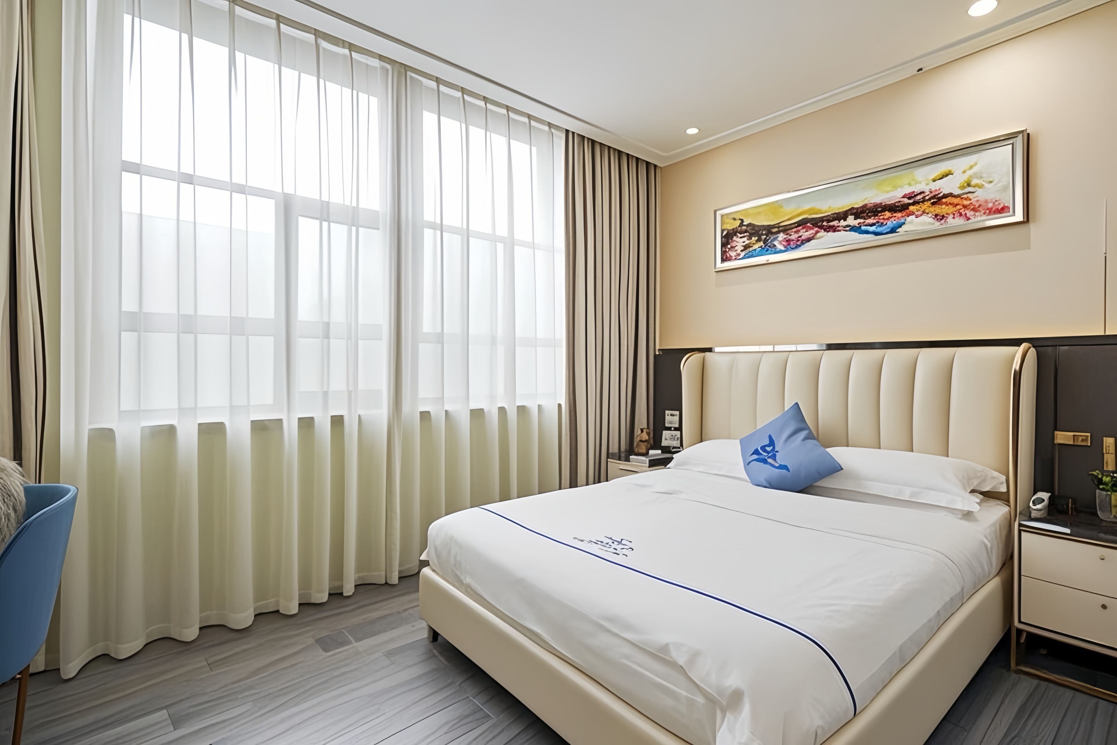 A hotel room with a large bed, a nightstand, a chair, a painting on the wall, and a window with curtains.