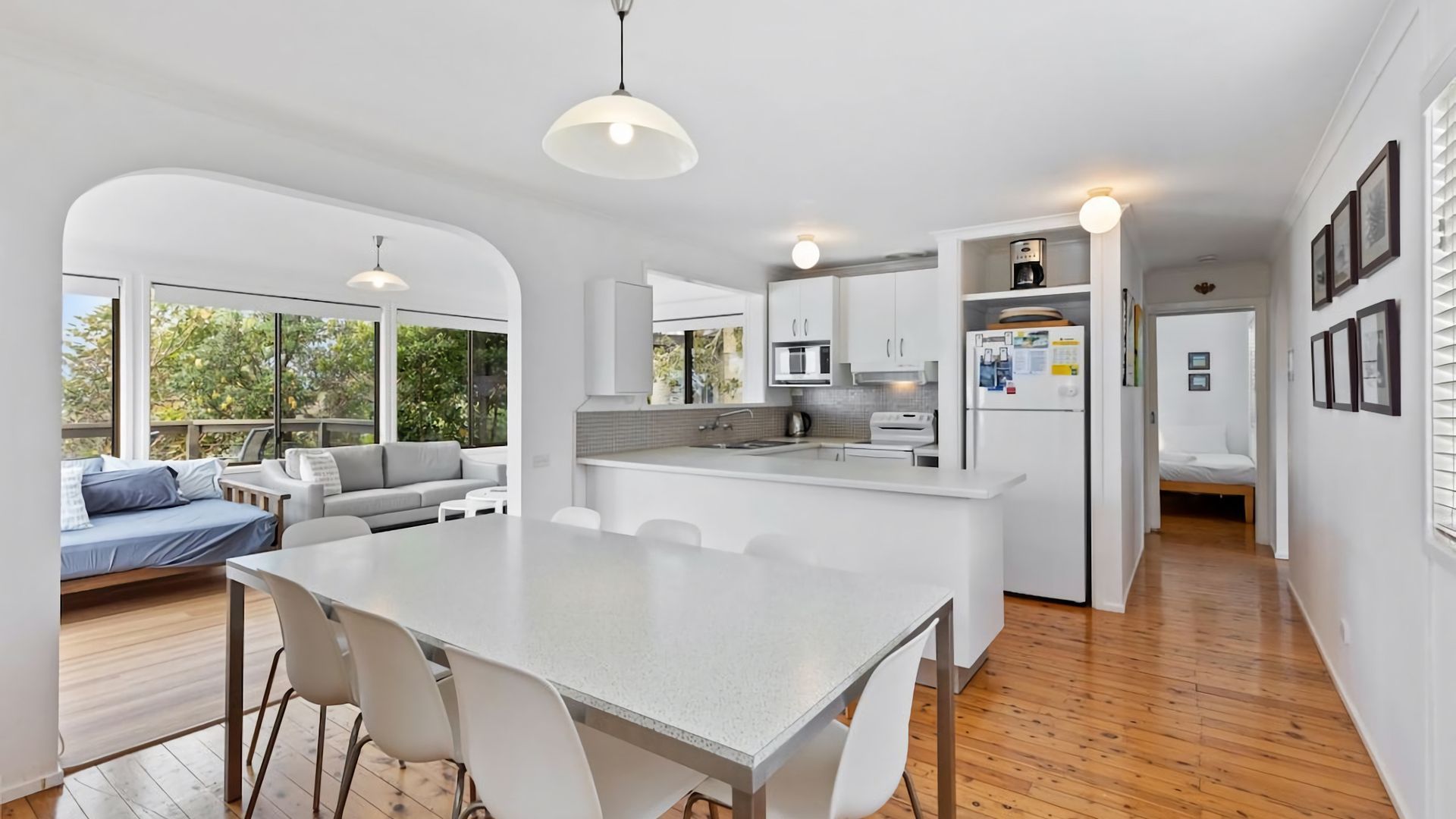 A modern kitchen with white cabinetry, a central island, and a dining area with a large table and chairs. There is a living area with a sofa and a view of the outdoors.