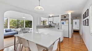 Una cucina moderna con mobili bianchi, un'isola centrale e una zona pranzo con un tavolo grande e sedie. C'è anche una zona soggiorno con un divano e vista all'esterno.