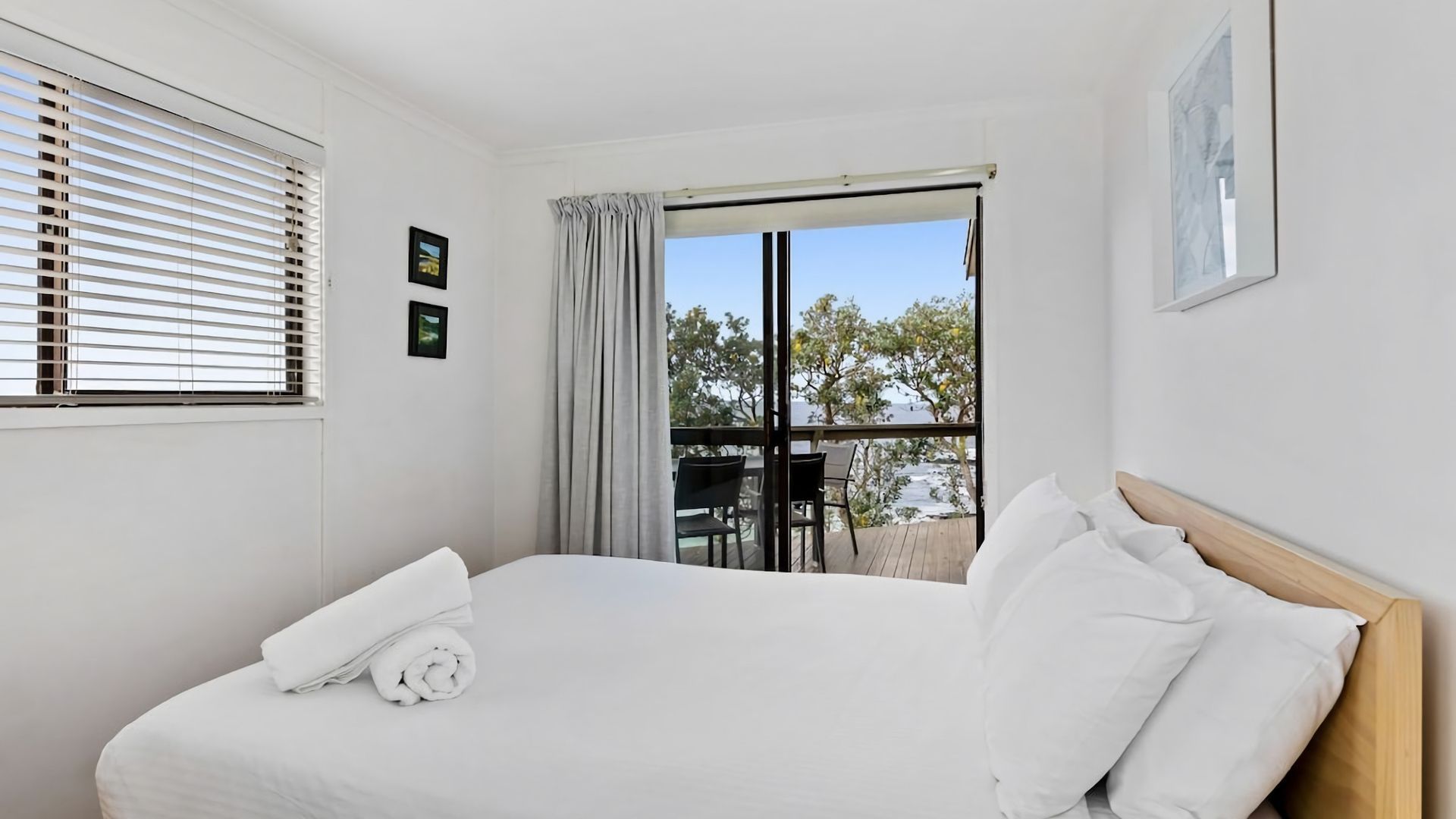 A hotel room with a bed, a window with blinds, a balcony with a table and chairs, and a view of trees and buildings.