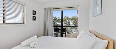 Une chambre d’hôtel avec un lit, une fenêtre avec des stores, un balcon avec une table et des chaises, et une vue sur les arbres et les bâtiments.