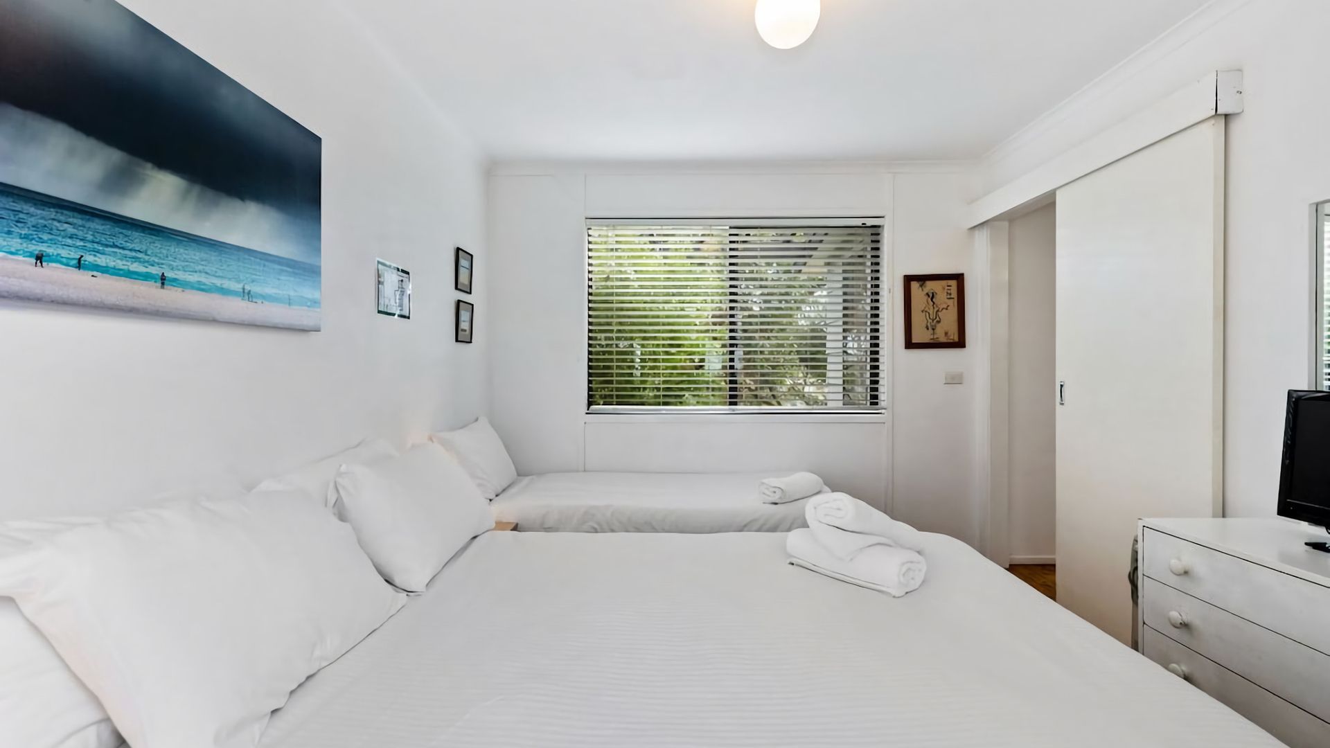 Habitación de hotel con dos camas, un ventanal con persianas, un cuadro en la pared y un televisor en una cómoda.