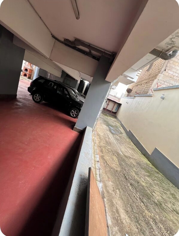 A parking garage with a red floor, a black car parked inside, and a view of a building with a brick facade outside. - Résidence Hibiscus (Yaoundé)
