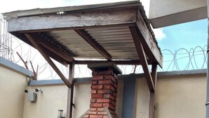 Un four extérieur en briques, surmonté d’un toit en métal, est adossé à un mur. On aperçoit, en arrière-plan, une clôture en fil de fer barbelé.