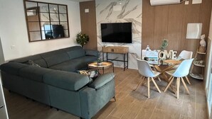 Un soggiorno moderno con un divano componibile, una televisione a schermo piatto, un'area pranzo con un tavolo in vetro e sedie, e un pannello decorativo a parete.