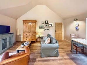 Un salon de style industriel, avec un plafond haut, un meuble en bois, un canapé gris, un fauteuil en cuir, une table à manger avec une chaise, et un téléviseur posé sur un meuble.