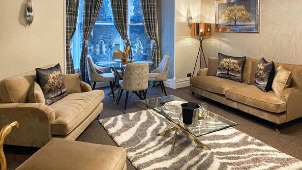 A living room with a sofa, armchair, coffee table, and a view of a building through large windows. - Merewyke Boutique Apartment - UK51055 (Bowness-on-Windermere)