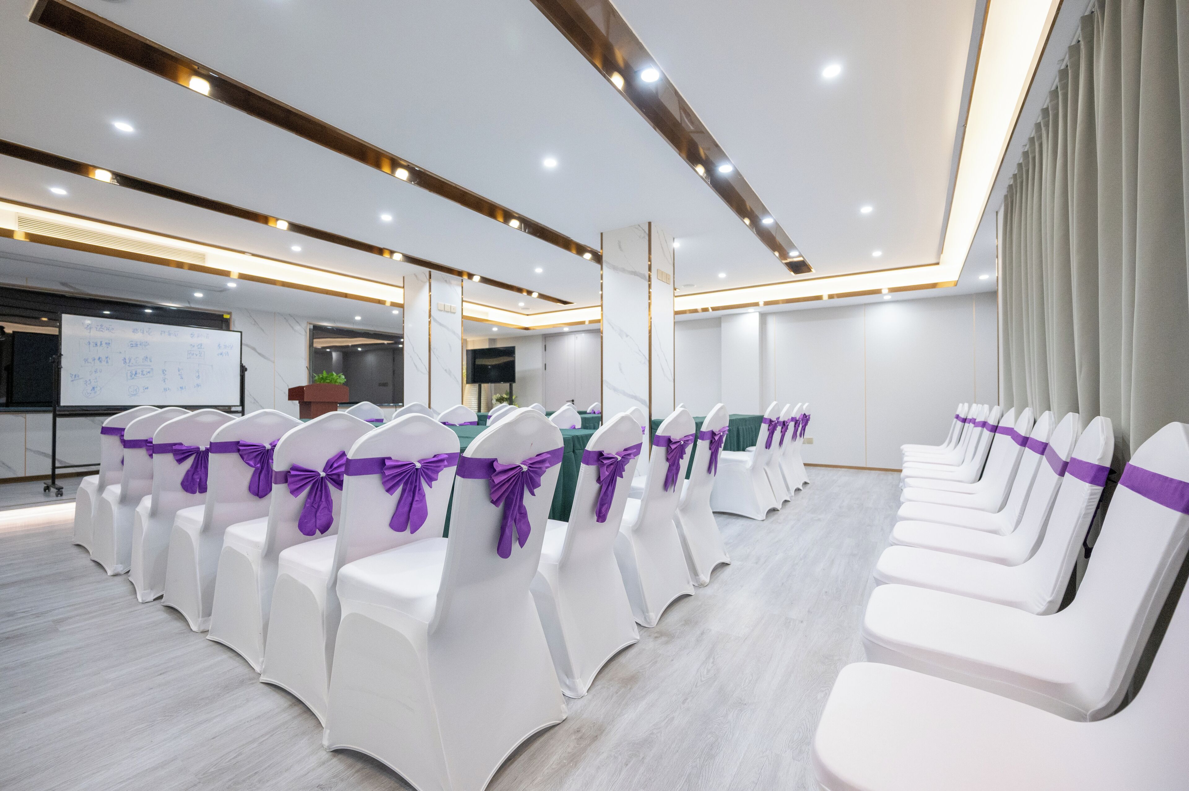 A conference room with rows of white chairs with purple sashes, a whiteboard, and a projector screen.