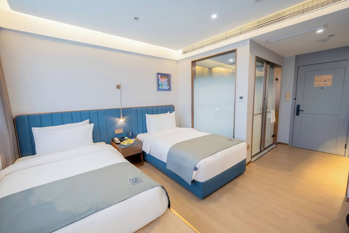 A hotel room with two beds, a nightstand with a lamp, a painting on the wall, and a door leading to a bathroom.