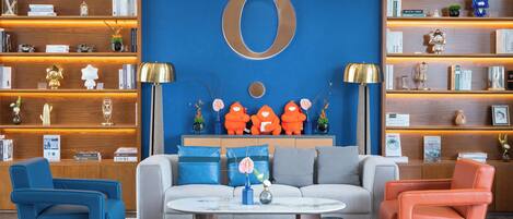 A modern living room with a large blue wall, a sofa, two armchairs, and a coffee table. There are shelves with books and decorative items, and a large 'O' on the wall.