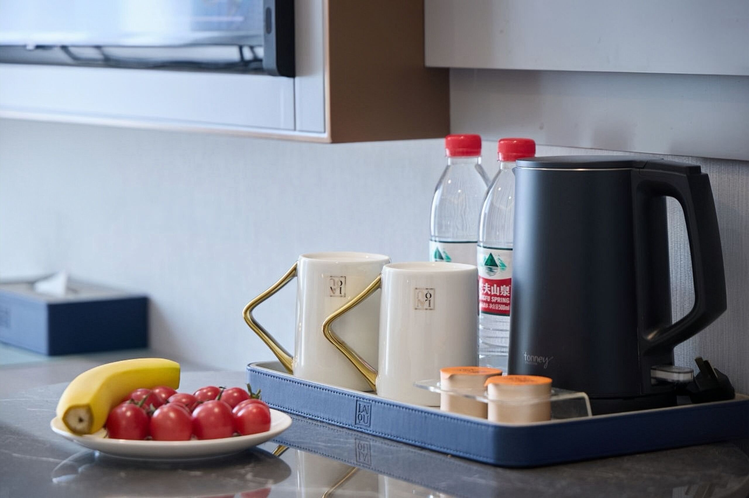 Una encimera de cocina con una bandeja azul que contiene una banana, tomates y un plato. Hay dos botellas de agua, una cafetera negra y dos tazas blancas con asas doradas.