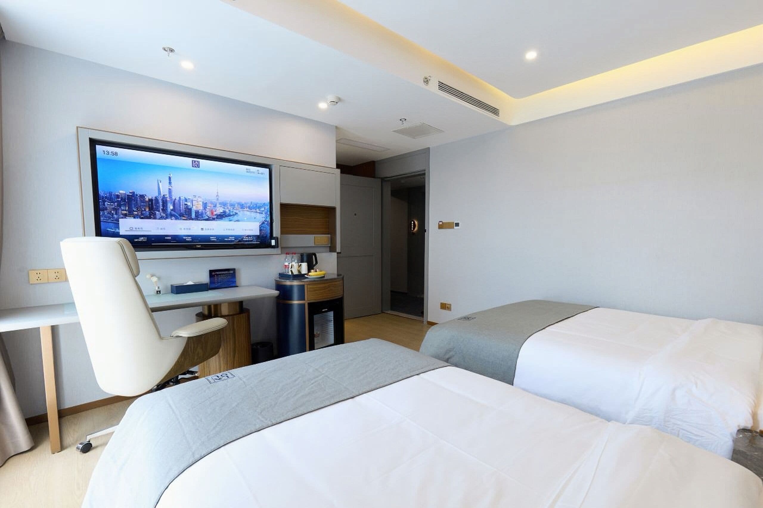 Ein Hotelzimmer mit zwei Betten, einem Schreibtisch mit Computer, einem Fernseher, der eine Stadtansicht zeigt, und einer Tür zu einem weiteren Raum.