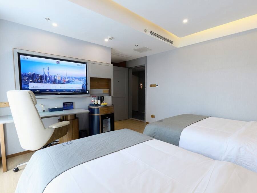 Ein Hotelzimmer mit zwei Betten, einem Schreibtisch mit Computer, einem Fernseher, der eine Stadtansicht zeigt, und einer Tür zu einem weiteren Raum.