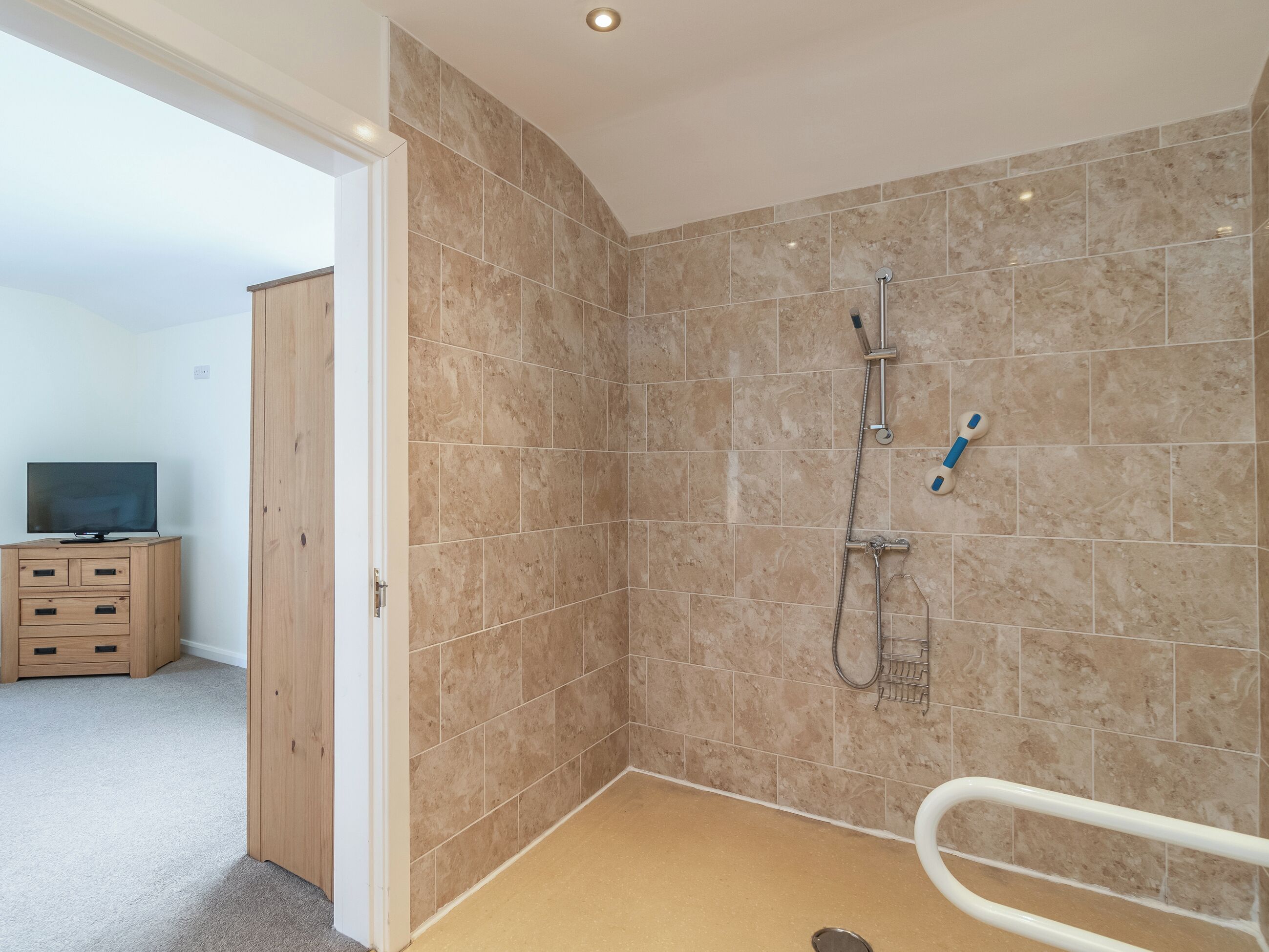 A bathroom with beige tiles, a shower, a toilet with a safety bar, and a wooden cabinet with a TV.