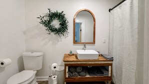 Un baño con inodoro blanco, tocador de madera con lavabo blanco, espejo, toallero y cortina de ducha.