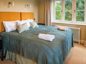 A neatly made bed with a green bedspread, white pillows, and a rolled white towel. There is a window with curtains, a radiator, and a wall-mounted lamp.