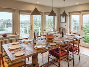 Un espace repas avec une longue table en bois, dressée pour six personnes, de grandes fenêtres et une vue sur la verdure environnante.