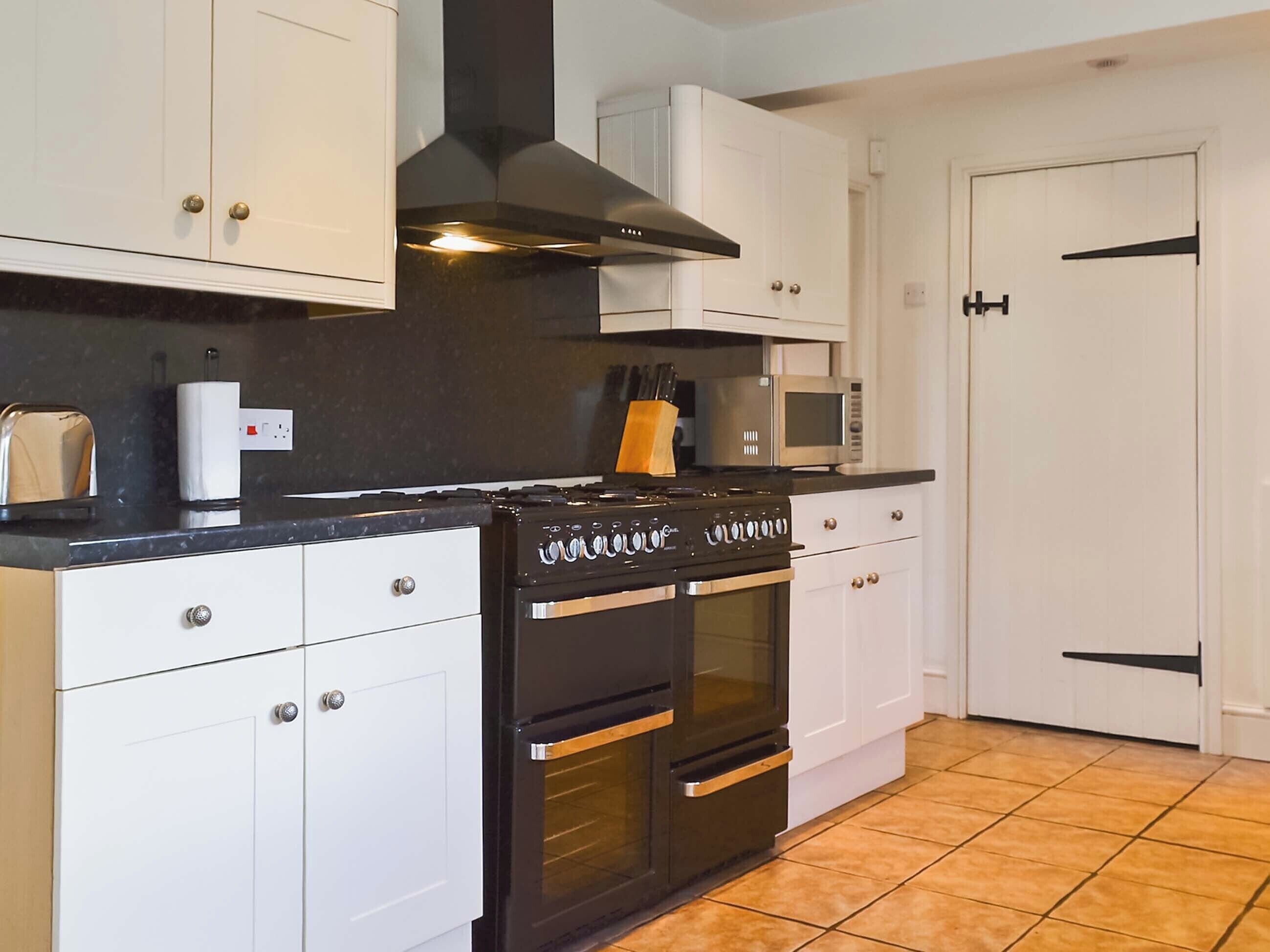 Une cuisine avec des meubles blancs, une cuisinière noire, un four à micro-ondes et un sol carrelé.