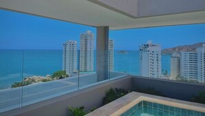 Una habitación de hotel moderna con un balcón amplio, vista al océano y edificios altos al fondo.