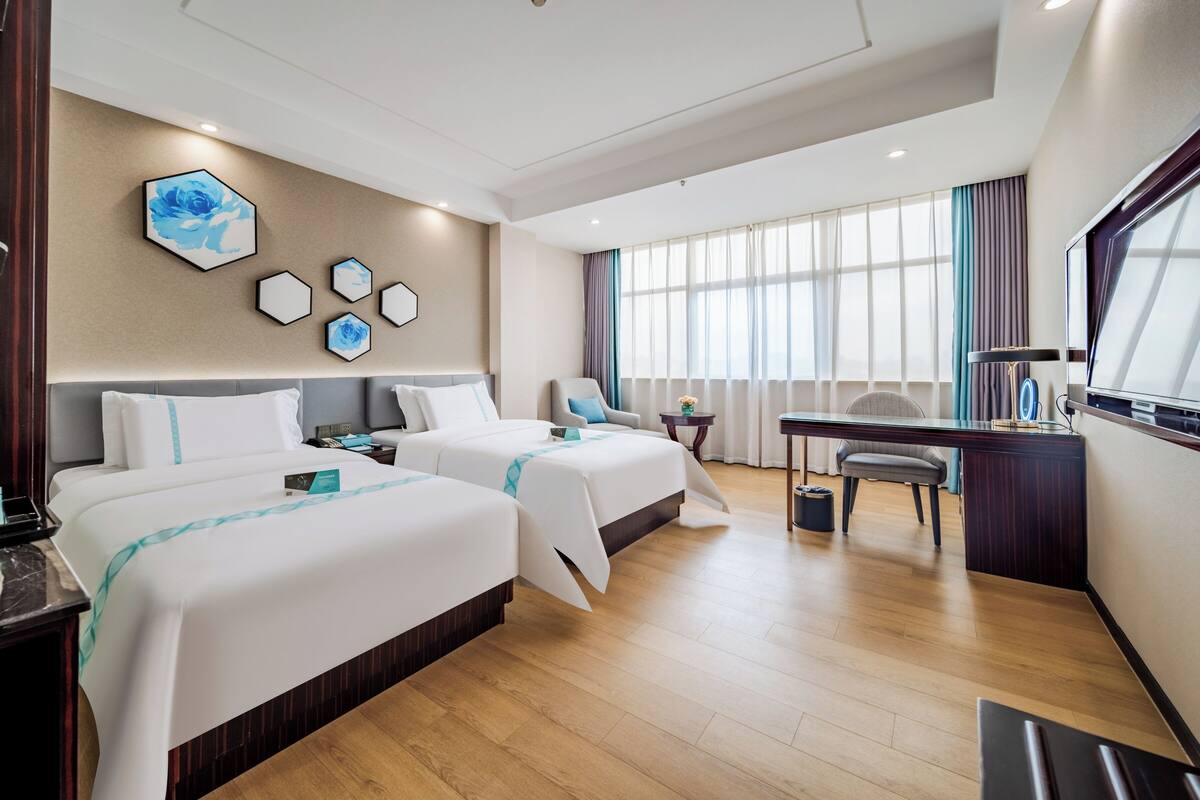 Ein Hotelzimmer mit zwei Betten, einem Schreibtisch, einem Stuhl und einem großen Fenster mit Vorhängen.