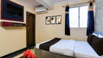 A hotel room with a bed, a TV, a small table with flowers, and a window with curtains.