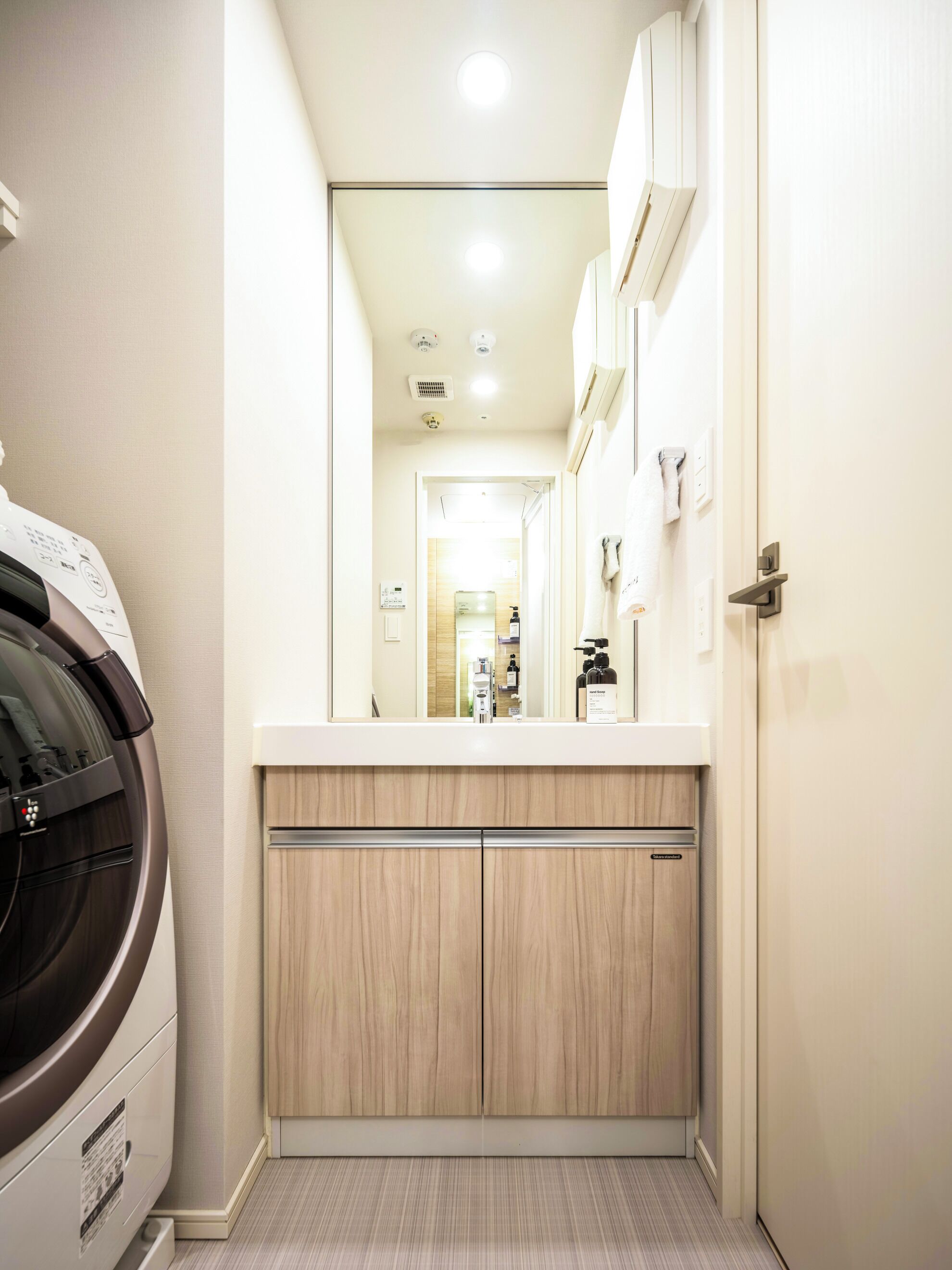 A compact laundry area with a washing machine, a countertop, and a mirror reflecting a bathroom with a sink and towel rack.