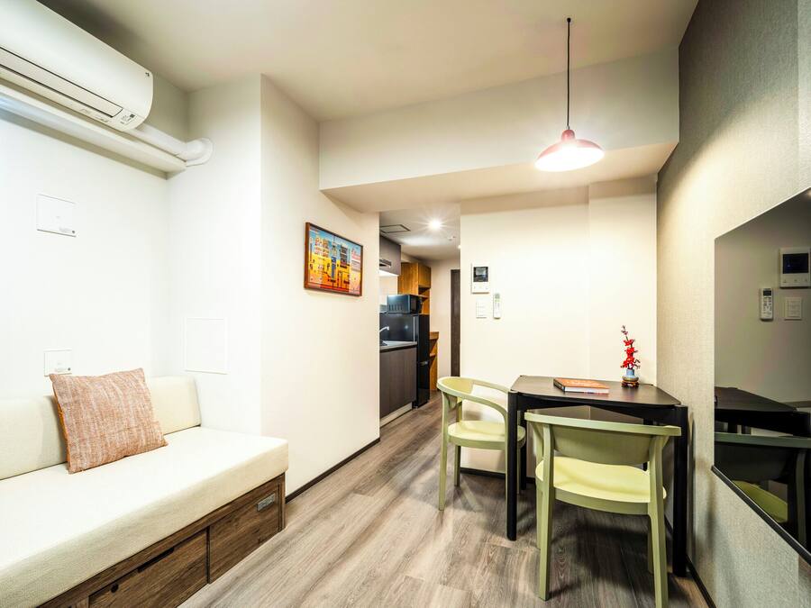 A modern, well-lit room with a sofa, a dining table with chairs, a small kitchen area, and a hanging light fixture.