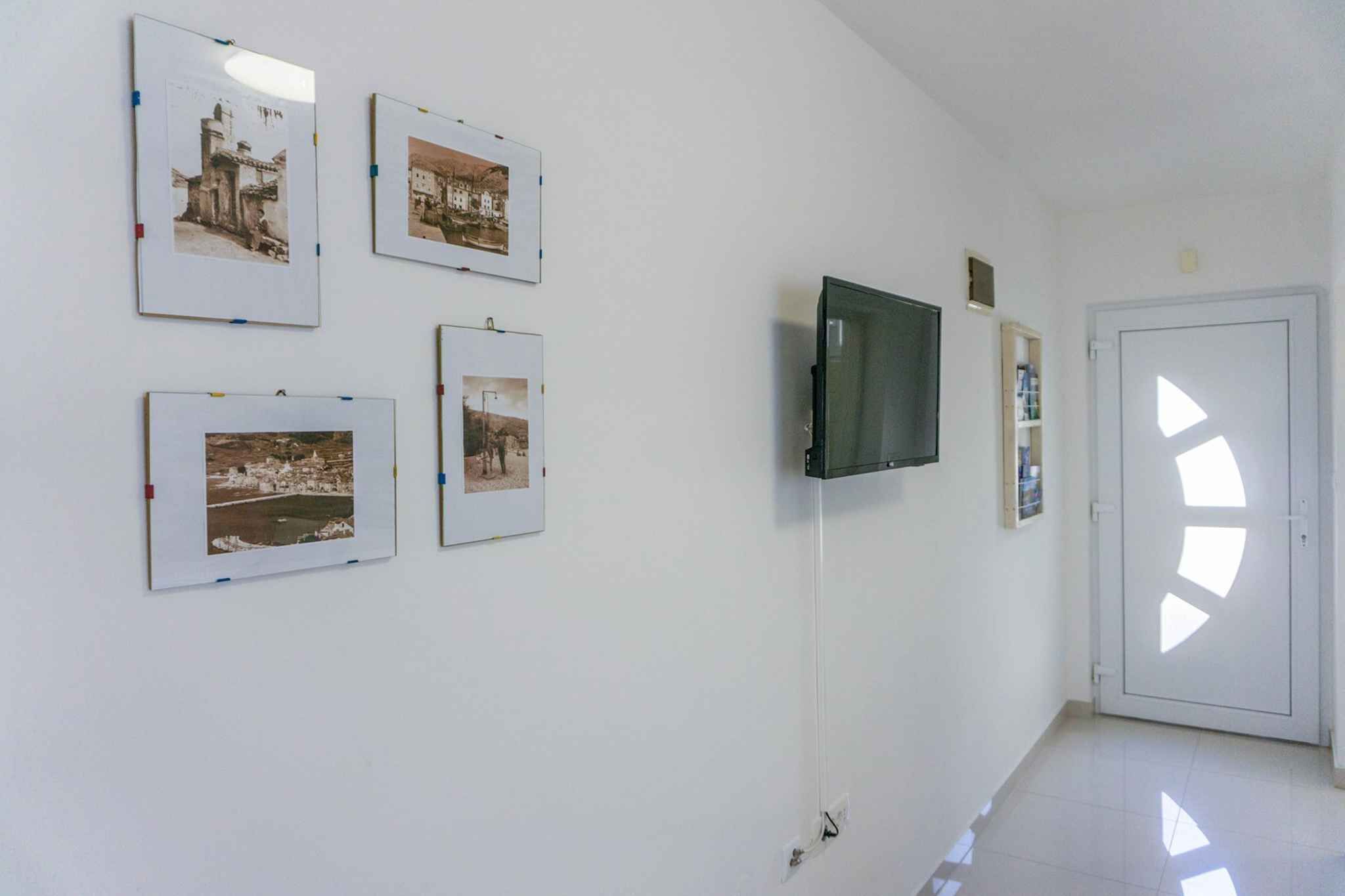 Een gang met ingelijste foto's aan de muur, een aan de muur bevestigde televisie en een deur met een geometrisch patroon.