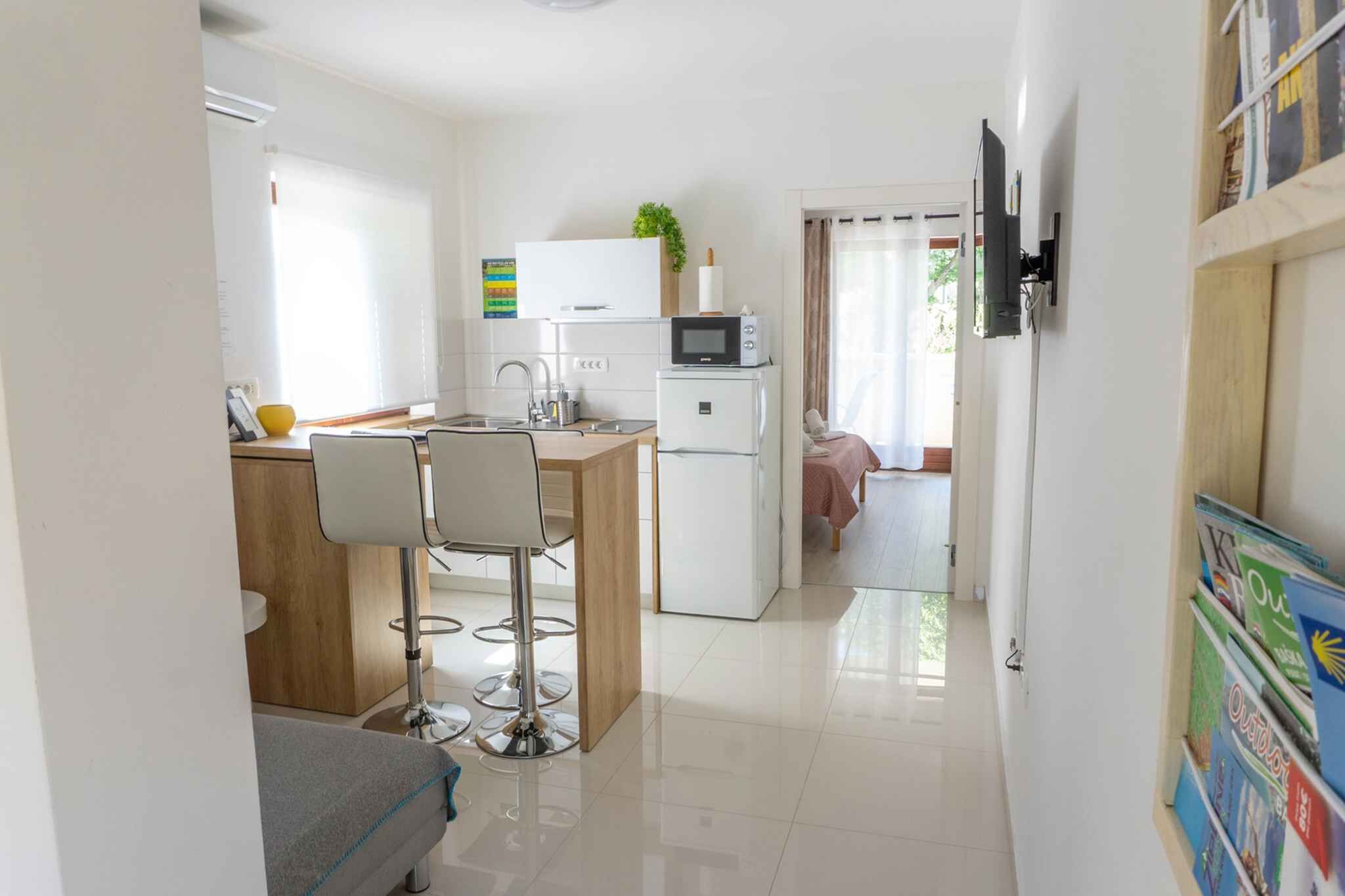 A compact kitchen with a microwave, sink, and a small refrigerator. There is a dining area with a table and chairs. A bed is visible in the background.