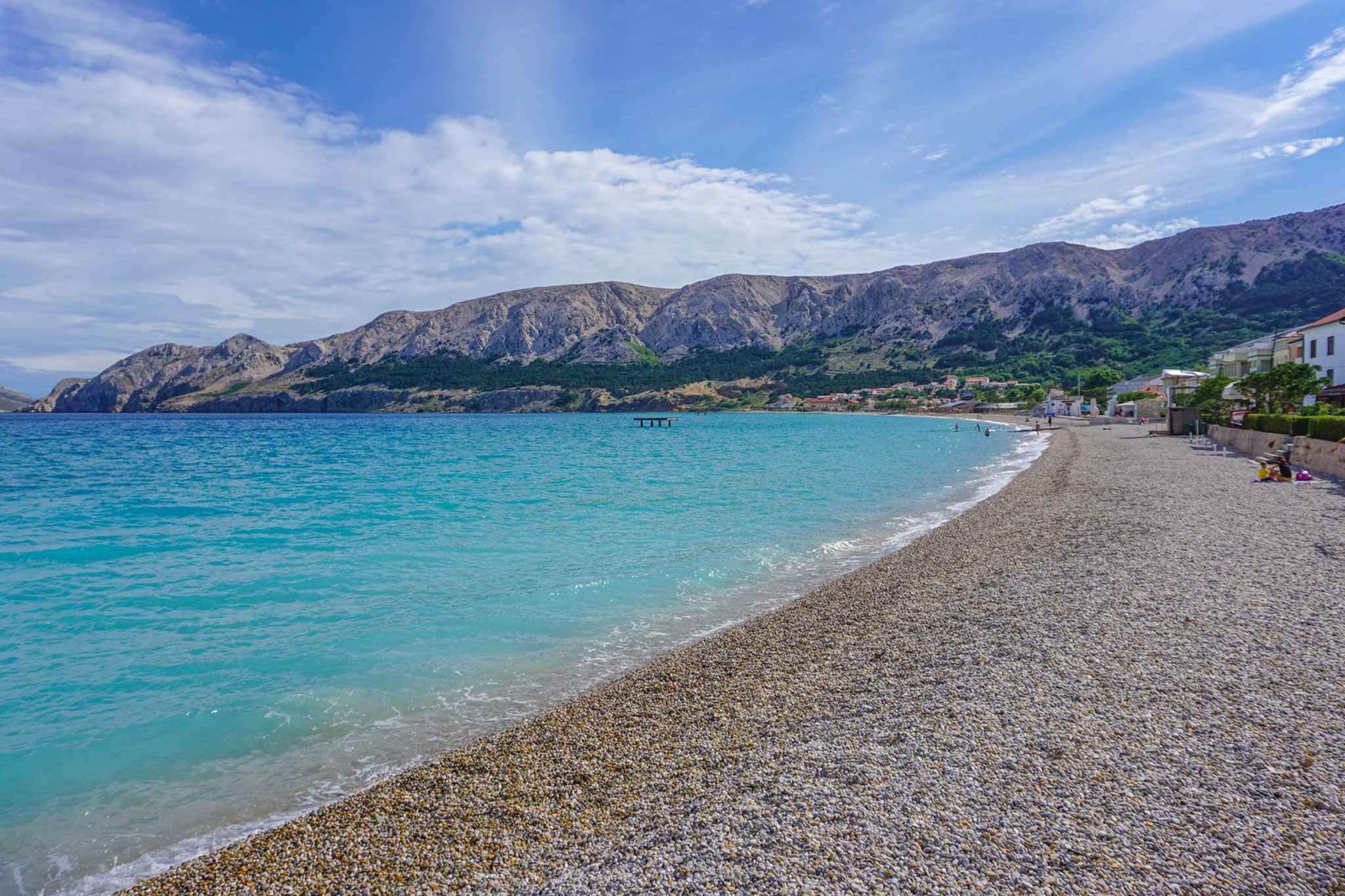 A pebble beach with clear turquoise water, a bench, and a mountain backdrop.