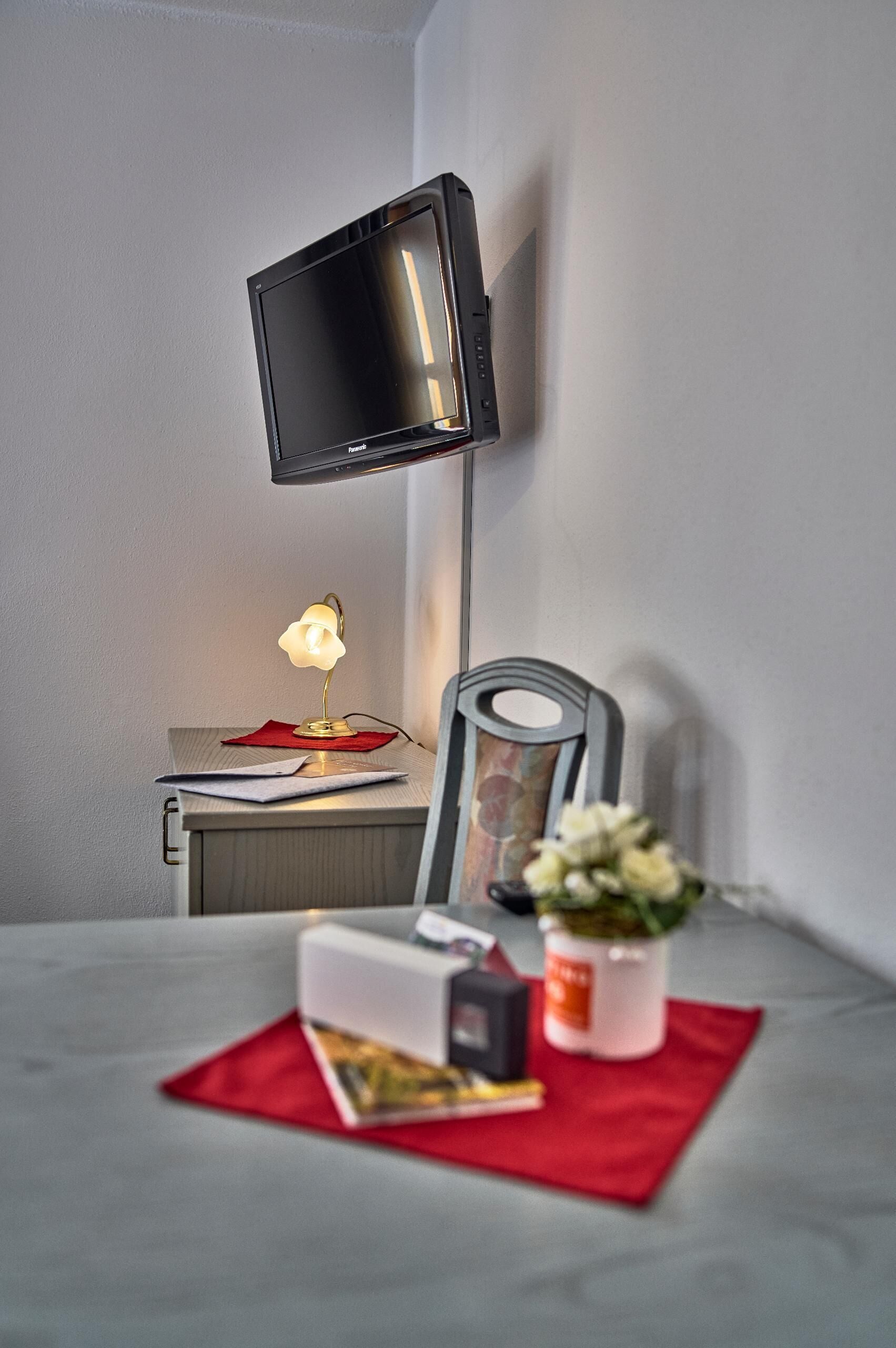Un espace de travail bien aménagé, avec un bureau, un téléviseur à écran plat fixé au mur, une lampe de bureau, une chaise et une petite plante en pot.